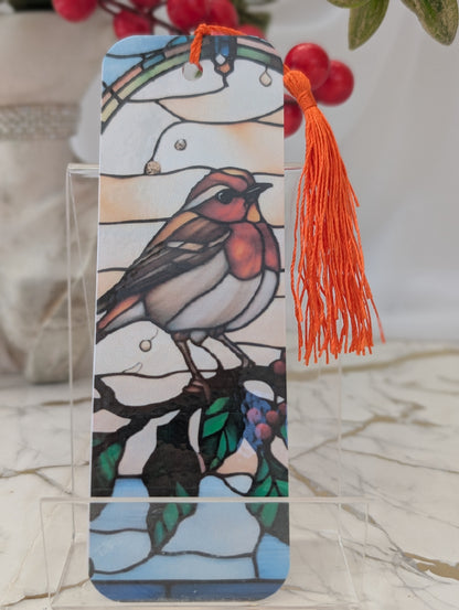 A stained-glass-Christmas Robin bookmark with an orange tassel, displayed on a clear acrylic stand. The bookmark features a robin perched on a branch with blue, green, and orange details against a light background.