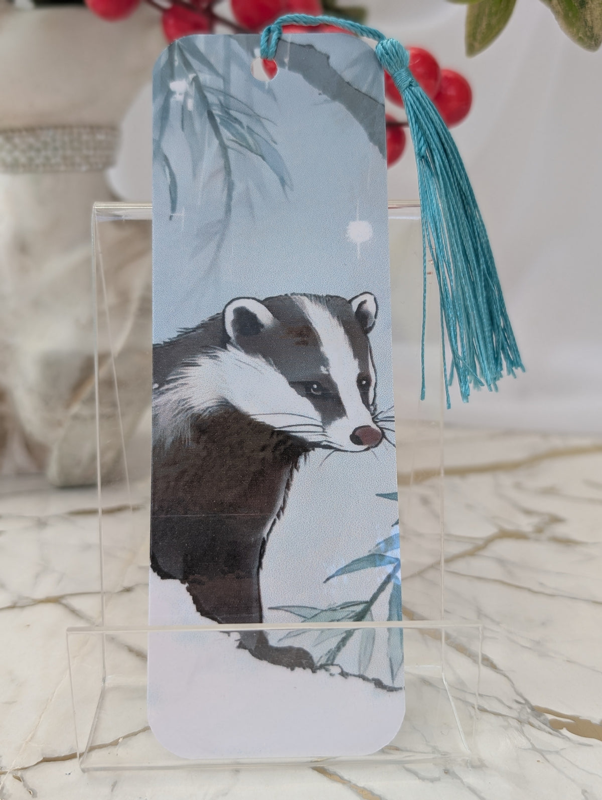 A rectangular paper bookmark featuring an illustration of a badger, displayed on a clear acrylic stand. The bookmark has a light blue background with a blue tassel hanging from the top.
