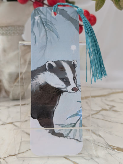 A rectangular paper bookmark featuring an illustration of a badger, displayed on a clear acrylic stand. The bookmark has a light blue background with a blue tassel hanging from the top.