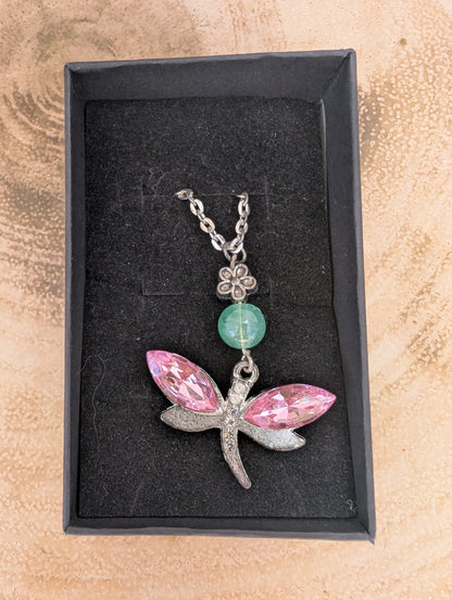 Silver tone dragonfly pendant with pink crystal stone wings and jade coloured bead and silver flower connector, in a black gift box on top of a wooden background