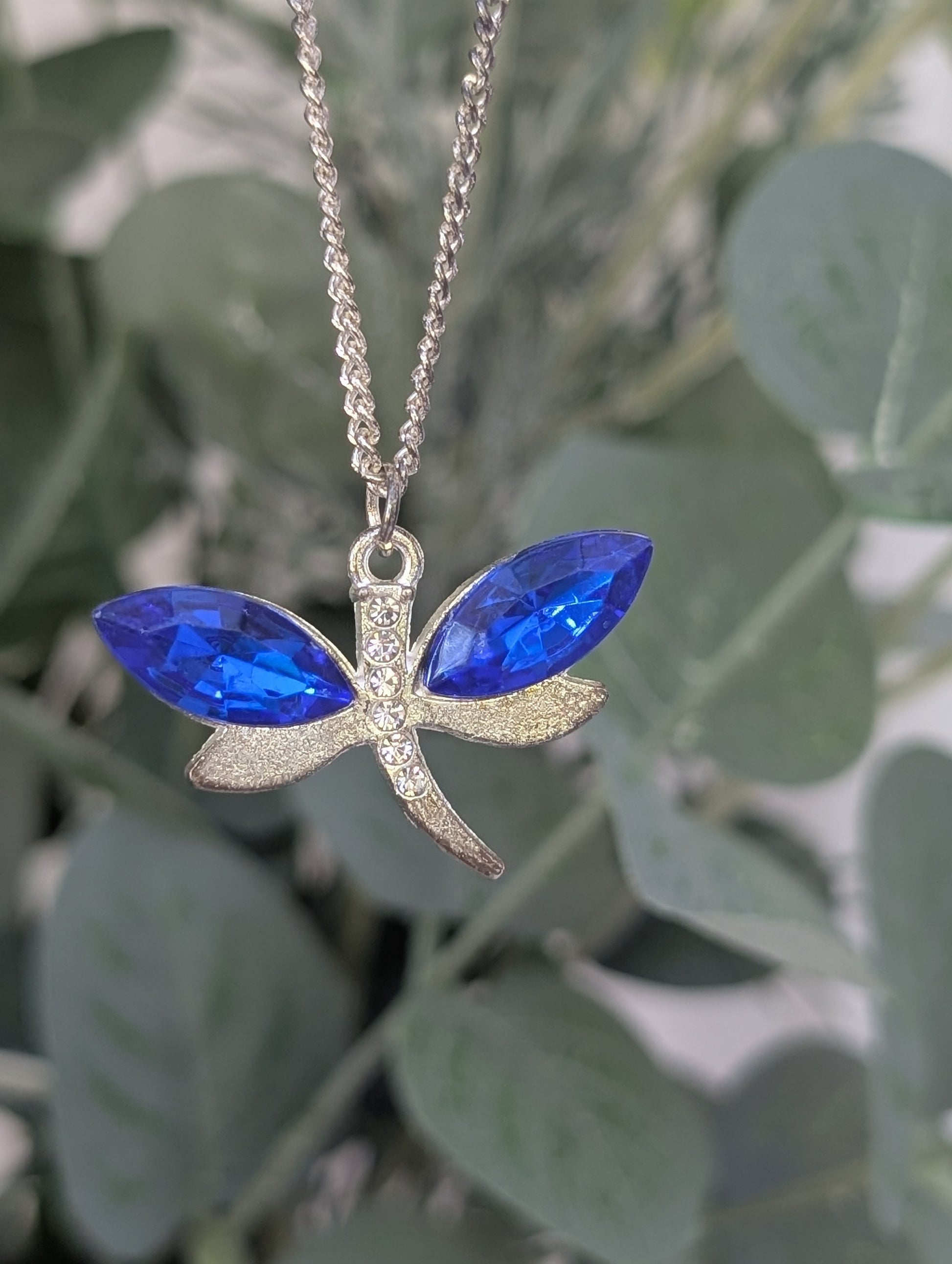 close up of silver tone dragonfly pendant wit sapphire blue coloured crystal stone wings and clear diamante body accents, against blurred green leaf background