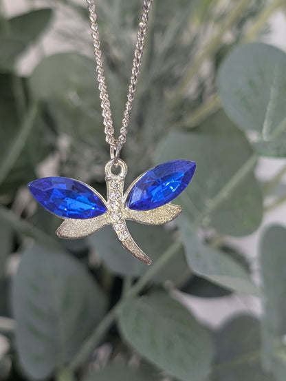 close up of silver tone dragonfly pendant wit sapphire blue coloured crystal stone wings and clear diamante body accents, against blurred green leaf background